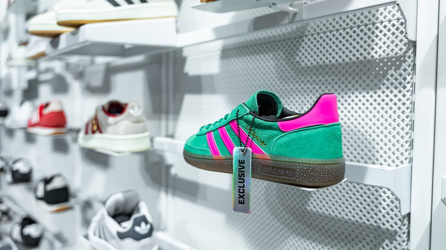 A green and bright purple pair of Adidas Samba shoes is sitting on a shelf in a sneaker store surrounded by several other Adidas shoes.