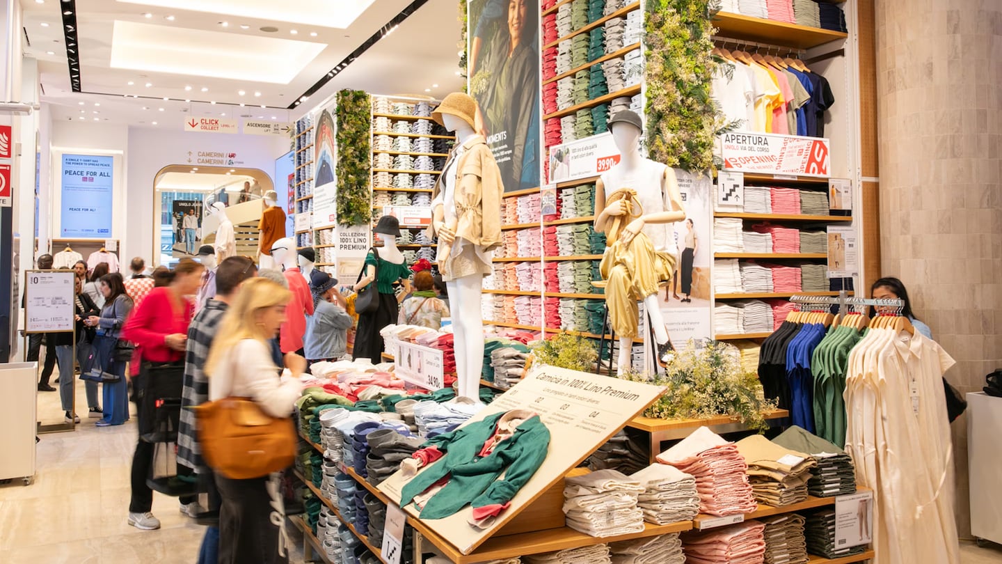 Figures shop in a Uniqlo store. To the right there are racks of clothes and shelves of different coloured ready-to-wear items. Customers sift through the offering to the left of the image. In the middle, mannequins stand displaying the clothes.