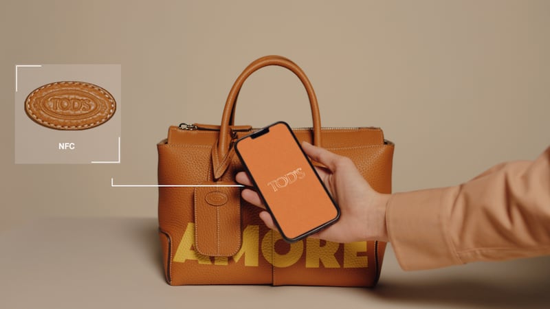 A hand reaches out with an iPhone to scan the logo of a Tod's Di Bag containing an NFC chip.