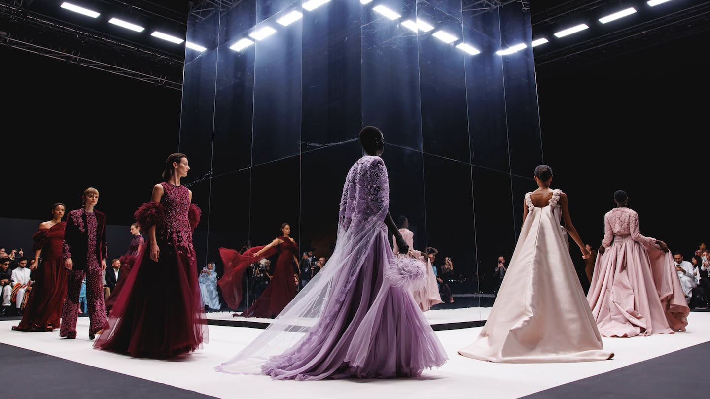 Models walk round a square runway, the central pillar made of mirrors, wearing dramatic evening-wear in purple hues.
